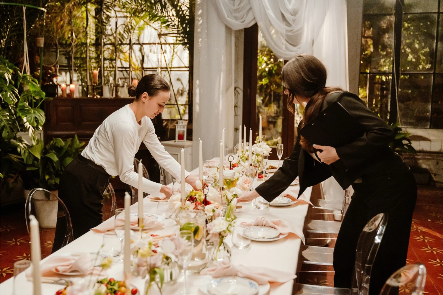 Two women setting a long banquet table with candles, florals, and blush napkins